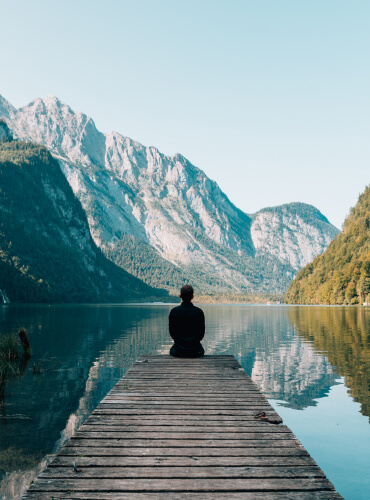 Foto mit einem Mann von hinten auf einem Steg an einem See umgeben von Bergen sitzend